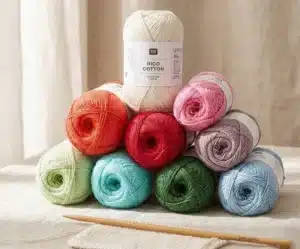 Photo professionnelle d'une pile de pelotes de fil Rico Cotton (100% coton mercerisé et gazé) en couleurs assorties (blanc, rouge, vert, bleu, rose, orange) reposant sur une nappe en lin. Une aiguille à tricoter en bois de 3 mm est posée au premier plan.