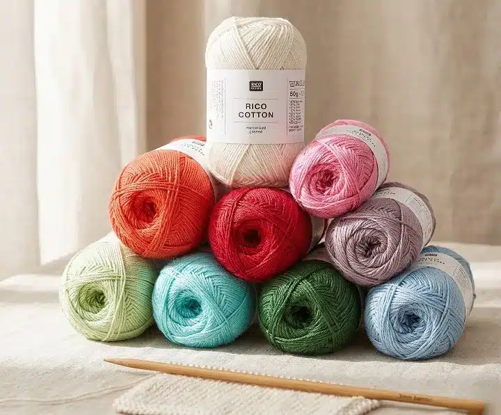 Photo professionnelle d'une pile de pelotes de fil Rico Cotton (100% coton mercerisé et gazé) en couleurs assorties (blanc, rouge, vert, bleu, rose, orange) reposant sur une nappe en lin. Une aiguille à tricoter en bois de 3 mm est posée au premier plan.